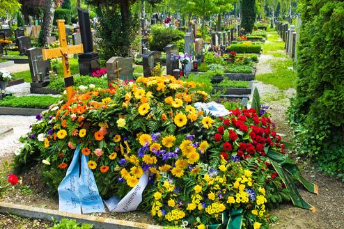 Bunt geschmücktes Grab mit einer Vielzahl von Blumen und einem Holzkreuz auf einem Friedhof umgeben von weiteren Gräbern.