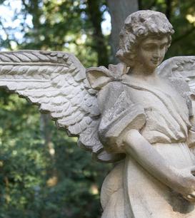 Skulptur eines Engels mit gefalteten Händen und großen Flügeln, umgeben von grünem Wald.