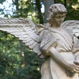 Skulptur eines Engels mit gefalteten Händen und großen Flügeln, umgeben von grünem Wald.