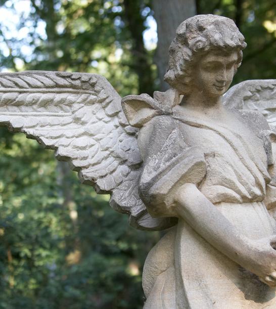 Skulptur eines Engels mit gefalteten Händen und großen Flügeln, umgeben von grünem Wald.